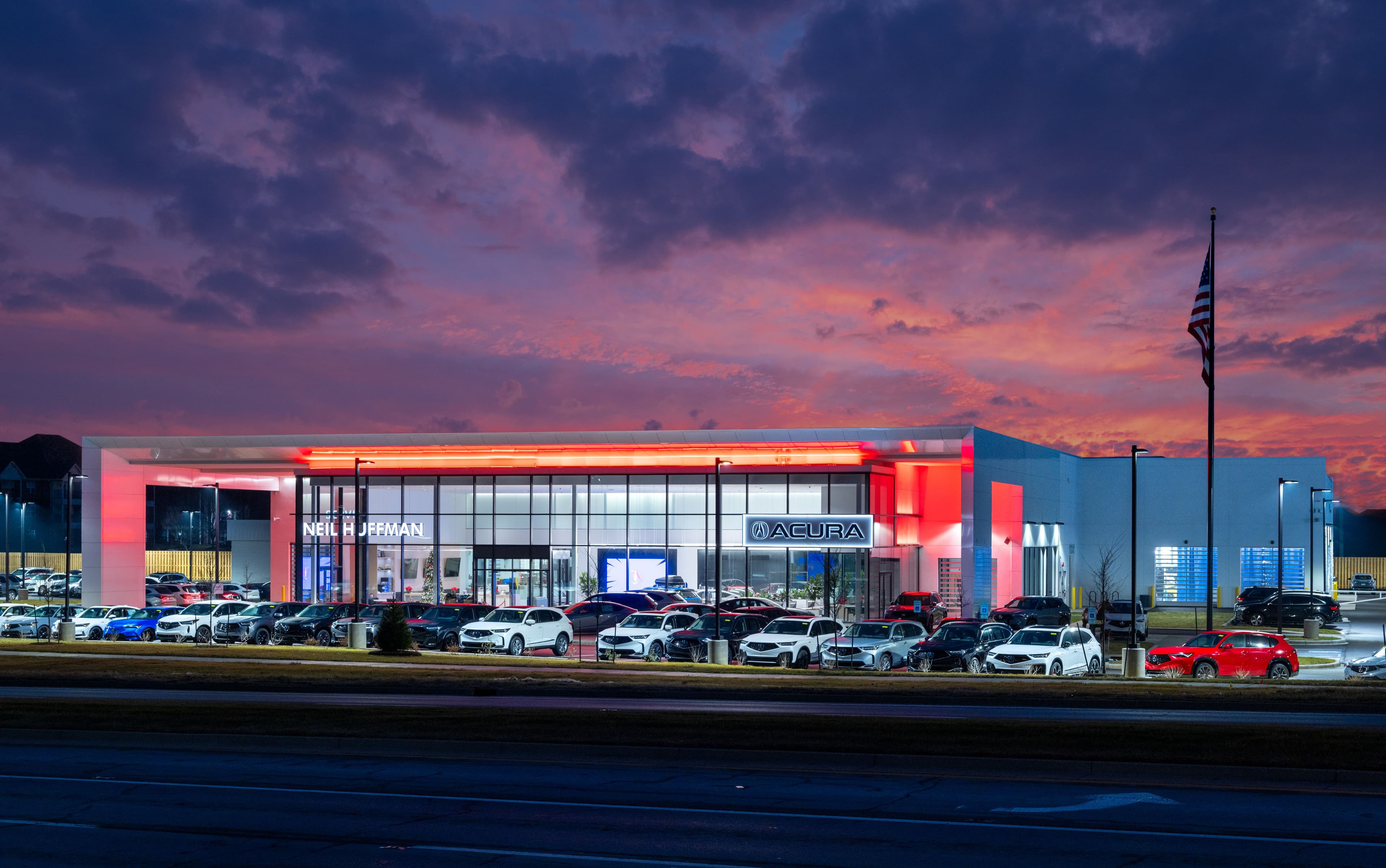 Exterior view of Neil Huffman Acura in Louisville, KY, showcasing the completed dealership at twilight with illuminated showroom windows, red architectural lighting, vehicle display lot, and dramatic sunset sky.