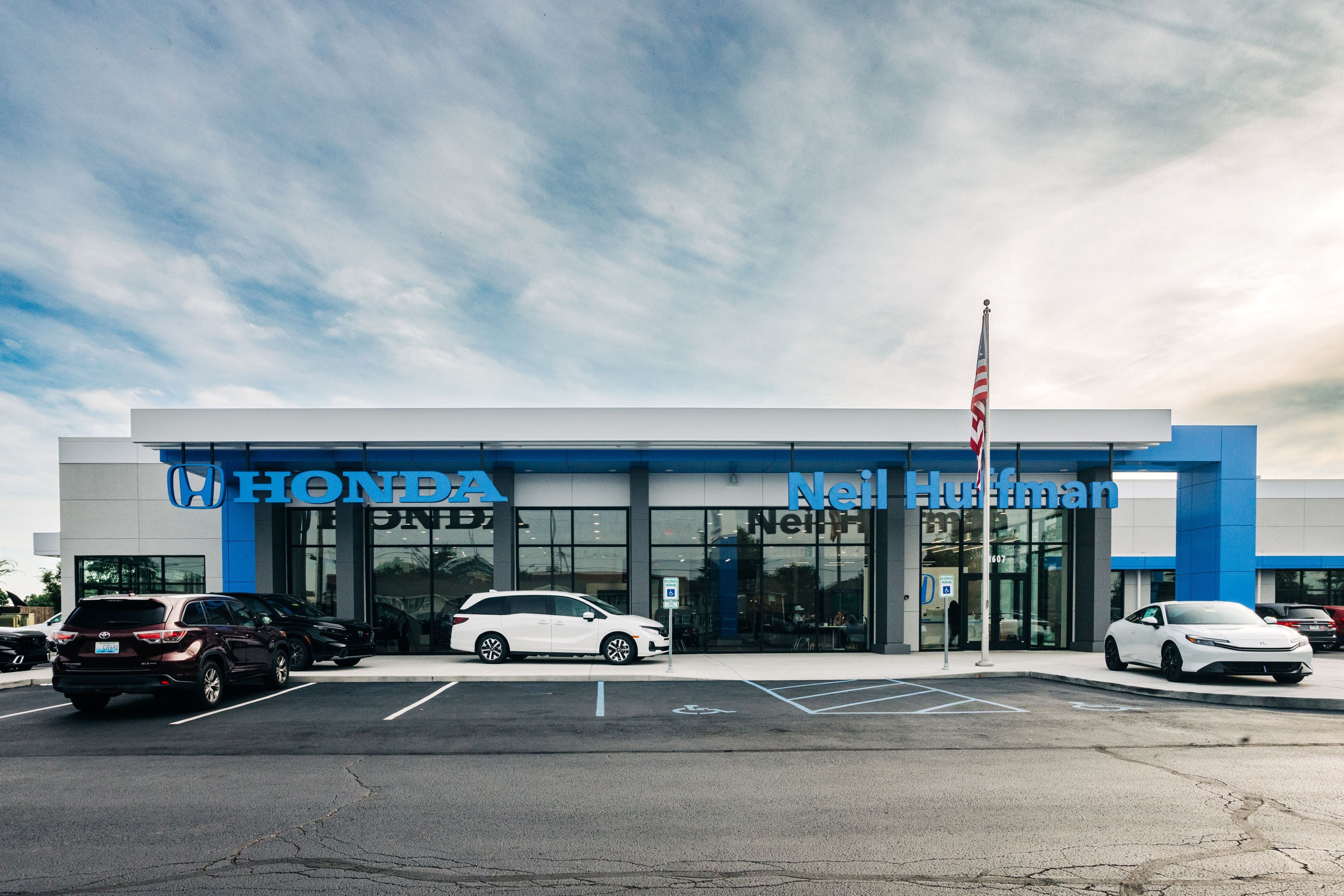 Exterior of the newly completed Neil Huffman Honda dealership, featuring a modern gray-and-blue facade, large Honda and Neil Huffman signage, front parking area, service bay entrance, and an American flag under an overcast sky.